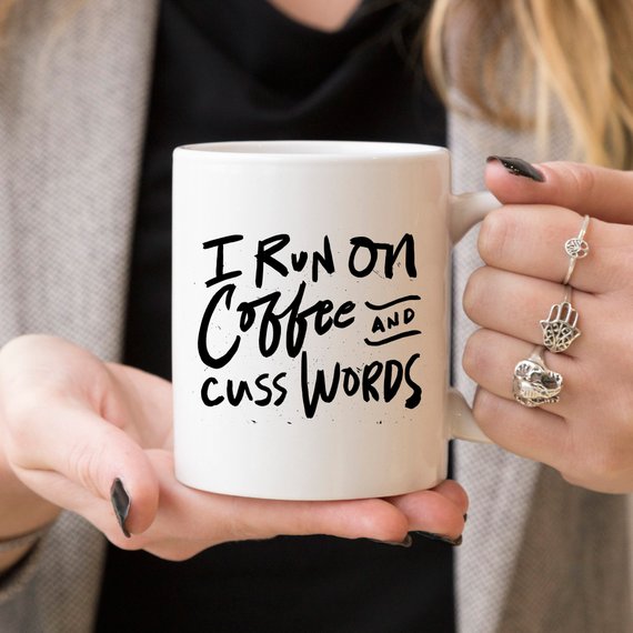 Ceramic coffee mug with "I Run On Coffee and Cuss Words" phrase held by a person.